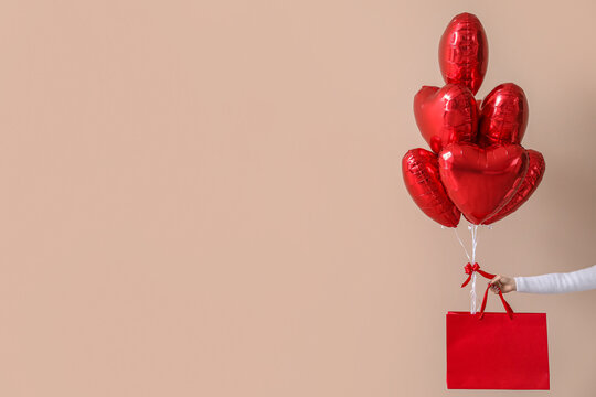 Woman with paper bag and heart-shaped balloons for Valentine's Day near beige wall