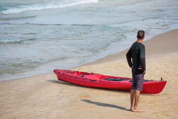 A man stands near a red kayak on a sandy beach.