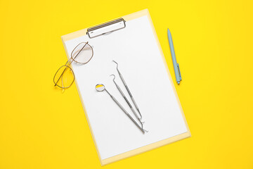 Clipboard with dental tools, eyeglasses and pen on yellow background