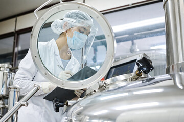 Female worker in protective wear writing equipment indicators in the card while working with...