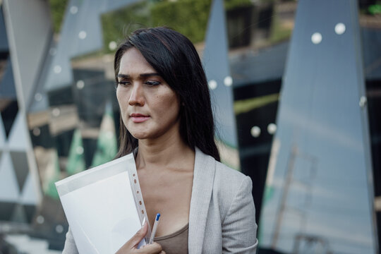 Thai woman with concerned look holding documents outside office building. Portrait of worried transgender person. Sex discrimination at work, diversity and social inclusion concepts