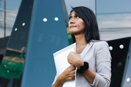 Thai woman with uncertain look holding documents outside glass office building. Doubtful transgender person in urban city. Job interview, college exams, diversity and social inclusion concepts