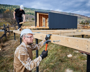Man worker building wooden frame house on pile foundation. Carpenter hammering bolt into wooden...