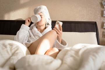 A woman sits on a bed wearing a white dressing gown, with a towel covering her head and drinks tea, while using a phone