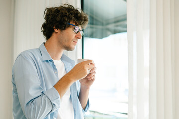 Young man standing near window thinking about work during coffee break