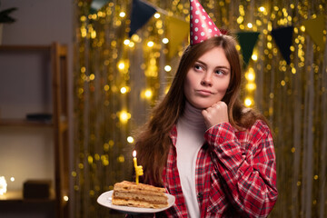 Obraz na płótnie Canvas Thinking young woman wearing party hat looking upward with her hand on her face and holding piece of cake while celebrate birthday party at home
