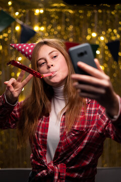Portrait of a happy young girl celebrating her birthday party at home, taking a selfie with festive horn