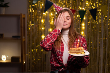 Portrait of happy young woman closes eyes and smiles, makes face palm. Overjoyed female holds birthday cake as present, wears casual clothes on party at home