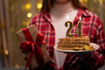 Young woman holding plate with tasty birthday cake and gift against defocused lights. 24th birthday.