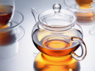 A transparent teapot with black tea and two cups on the sides on a mirrored table with reflections. Through the light. Studio shot. Horizontal.