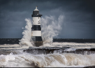 Lighthouse, black and white striped light house with big wave crashing over against a stormy sky. 