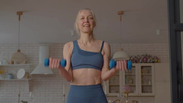 Active motivated attractive sporty fit senior woman doing dumbbell bicep curls exercise, strengthening upper arm muscles while practicing weight training in domestic room.