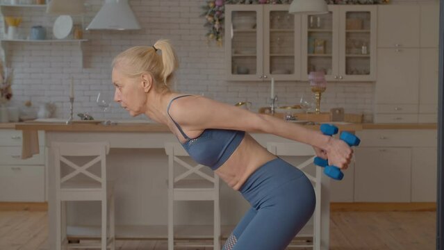 Active motivated pretty fitness senior woman in sportswear practicing dumbbell triceps kickback exercise, toning arms, increasing physical strength while doing weight training indoors.