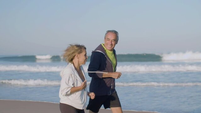 Cheerful mature couple running along ocean coast in morning. Side view of happy Caucasian man and woman in sportswear enjoying time together, doing physical activity and talking. Love, sport concept