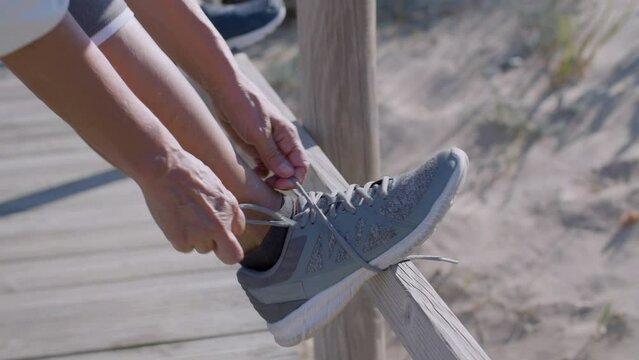 Unrecognisable mature woman lacing up sneakers on summer day. Close-up shot of athlete preparing for training, running or exercising on wooden path outdoor. Preparation, sport concept