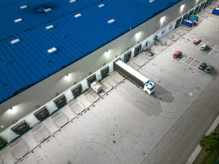 Aerial view of goods warehouse at Night. Logistics center in industrial city zone from above. Aerial view of trucks loading at logistic center. View from drone.
