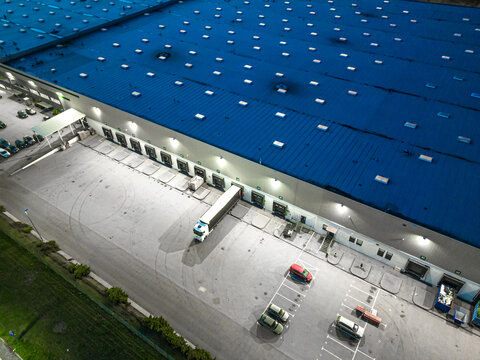 Aerial View Of Goods Warehouse At Night. Logistics Center In Industrial City Zone From Above. Aerial View Of Trucks Loading At Logistic Center. View From Drone.