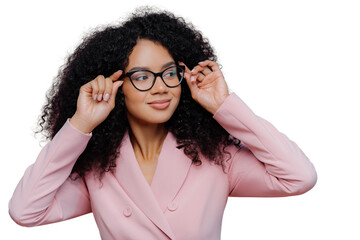 Headshot of attentive female boss keeps hands on frame of glasses, looks pensively away, wears rosy...