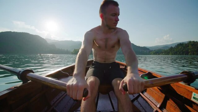 Young man with bare torso rows paddles in wooden boat on Bled lake sailing to island against high mountains at sunset in Slovenia