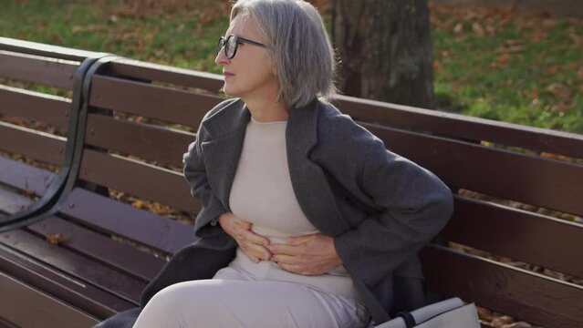 Elegant senior woman frowning in pain sitting on bench in park, feeling acute abdominal pain, suffering gastritis or duodenal ulcer, risk of ruptured appendix or congestion in the gallbladder