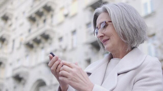 Smiling senior woman scrolling web pages on smartphone standing outdoor near big building, looking for apartment to rent in online application on smartphone, real estate business