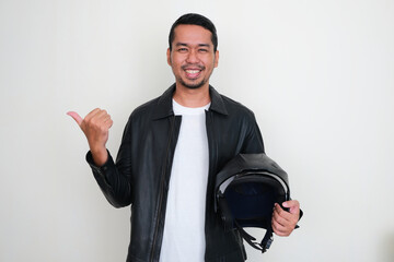 Adult Asian man wearing leather jacket holding motorcycle helmet smiling and pointing to the right...