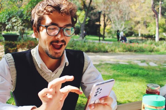 portrait of young man with glasses looking at phone in spring outfit in a green grass park 