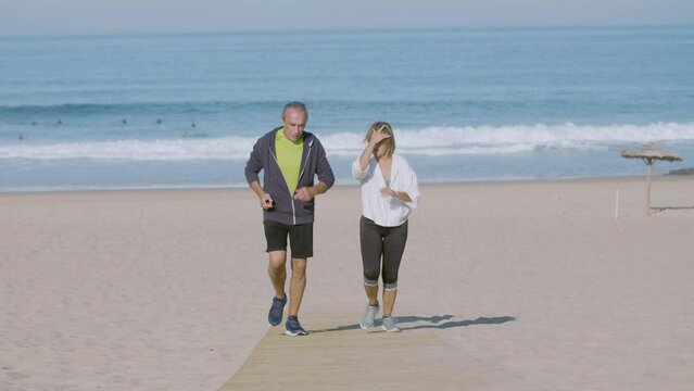 Cheerful Mature Couple Running Uphill On Sandy Beach. Front View Of Beautiful Man And Woman Jogging Together On Summer Day, Talking And Spending Time Together. Sport, Family Concept