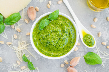 Traditional Italian basil pesto sauce in a white bowl with ingredients for cooking on a gray concrete background. Copy space.
