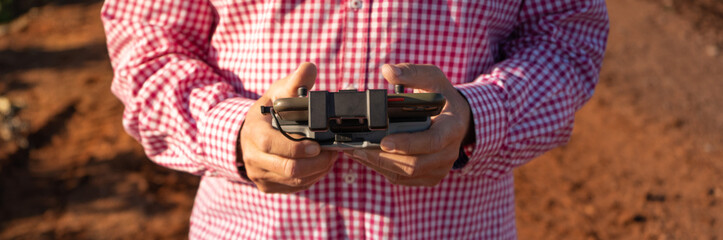 Drone control pad with a smartphone on a stand being used by the hands of an older farmer man. He wears a checkered shirt. Farmland background. Crop control
