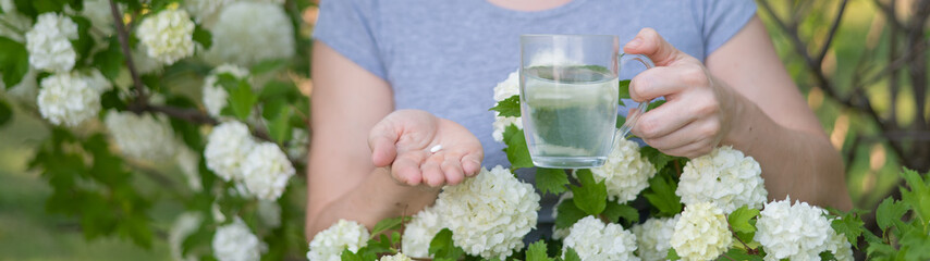 Woman holding an antihistamine tablet and a glass of water while in a blooming garden. 