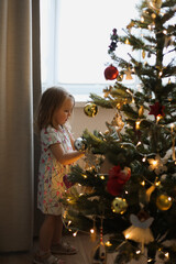 Little girl decorating christmas tree with toys and baubles. Cute kid preparing home for xmas celebration.