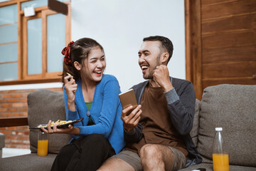 asian girl and boyfriend laughing while using cell phone together while sitting on sofa at home