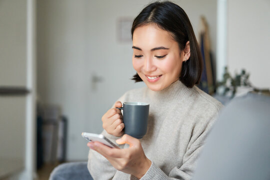 Portrait of smiling asian girl checking her news feed on smartphone and drinking coffee, sitting on sofa at home, browsing on mobile phone, reading