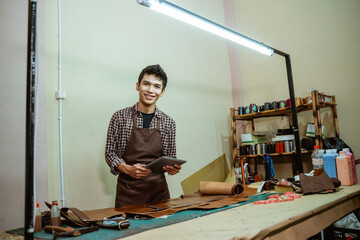 Young asian craftsman smiling at camera while using pad in leather workshop