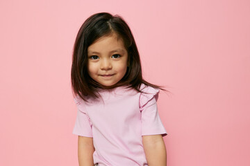 a little beautiful girl of preschool age stands on a pink background in a pink T-shirt with her hair down, looks at camera with incomprehension  - obrazy, fototapety, plakaty