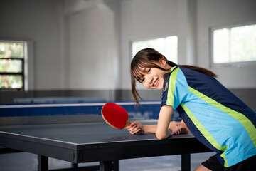 female athlete smiling while leaning on ping pong table and holding paddle in indoor building