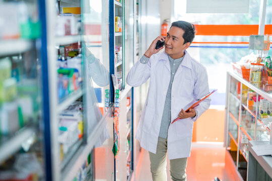male pharmacist in uniform making phone call while checking and holding clipboard in pharmacy