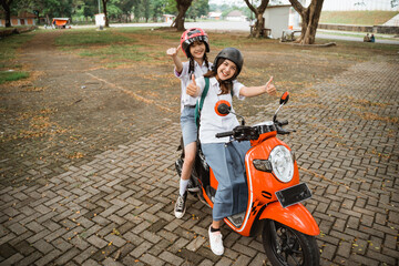 Two asian student girls wearing helmets with thumbs up riding a motorbike together