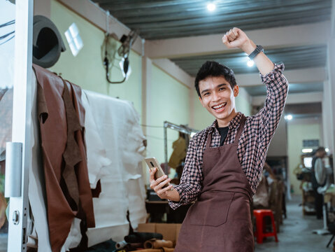 excited asian craftsman smiling using smartphone at leather craft workshop