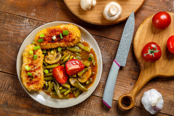 Green beans with mushrooms and cutlets sprinkled with green onions in a plate on a wooden table next to mushrooms, tomatoes and onions and a knife.