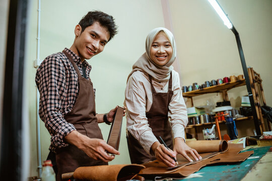 female craftsman in hijab with a male craftsman smiling while using a pencil and ruler measuring leather material