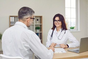 Friendly smiling young female doctor in a white coat, glasses and stethoscope sitting her at table with a laptop computer and giving a medical consultation to a mature man patient