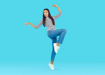 Happy young girl in casual outfit dancing in fashion studio. Full body shot of cheerful beautiful Caucasian woman in striped shirt and blue jeans dancing on blue colour studio background