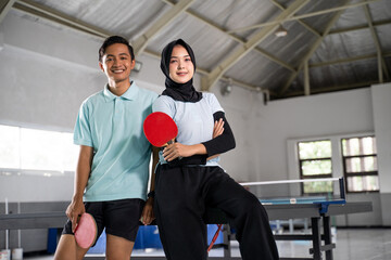 Fototapeta na wymiar man and woman athlete in hijab standing holding bat with crossed hands on ping pong table background