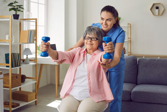 Smiling senior woman lifting dumbbells, therapist assisting her. Cheerful female nurse helping elderly patient to do physical exercise at home. Disabled, elderly people rehabilitation and support