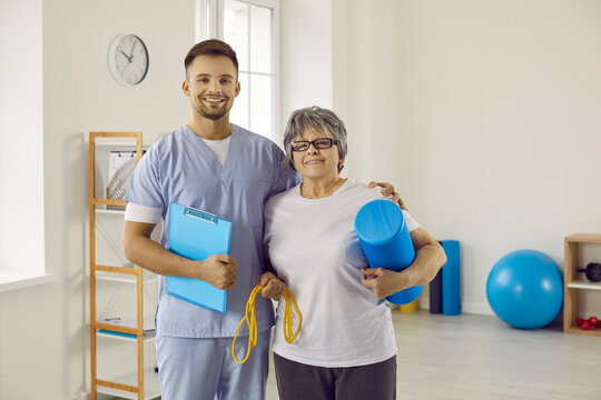 Portrait of happy physiotherapist and senior patient. Young worker with clipboard and old woman with sports equipment standing together in physiotherapy room. Health, exercise, rehabilitation concept - Powered by Adobe