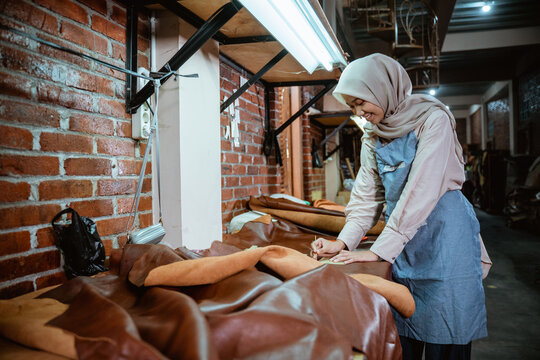 Female hijabi tailor in apron measuring leather material in leather workshop