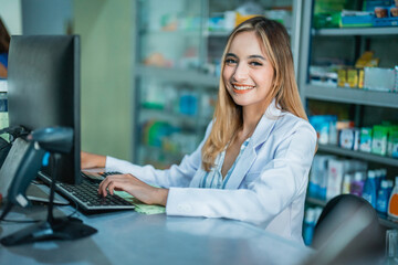 beautiful asian pharmacist in uniform smiling while working using pc computer in pharmacy