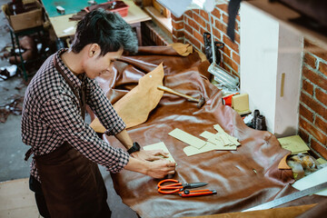 Fototapeta na wymiar Asian craftsman in apron working with leather clinging pattern paper in workshop